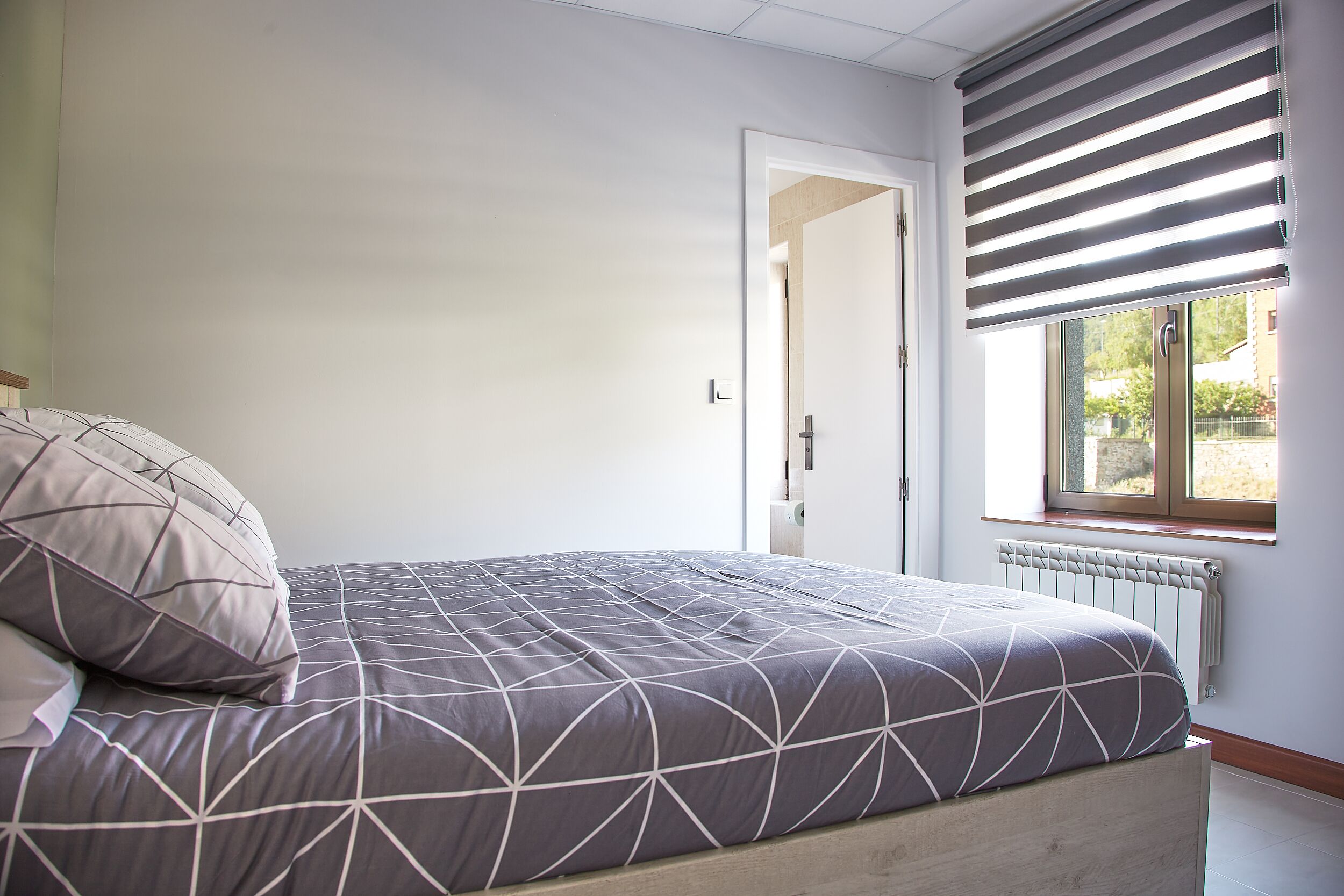 A bedroom with a bed, a radiator, a window with blinds, and a door leading to another room.