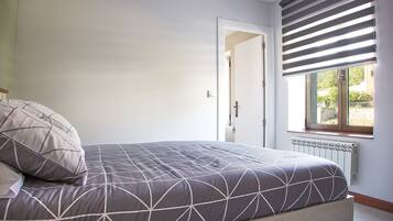 A bedroom with a bed, a radiator, a window with blinds, and a door leading to another room.