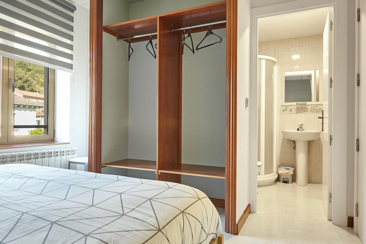Un dormitorio con cama, un armario de madera con perchas y un baño con lavamanos y espejo.
