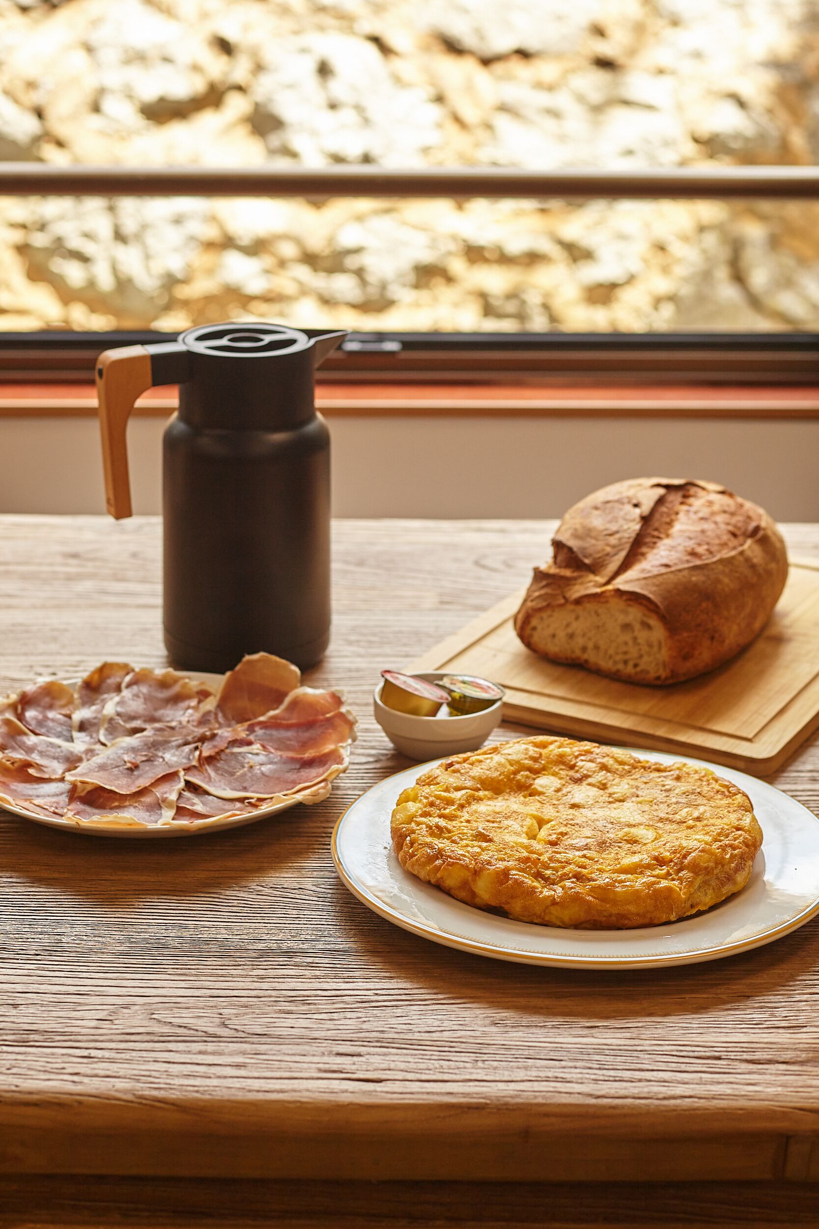 A wooden table with a loaf of bread, a plate of cured meats, a thermos, and a small bowl of olives.