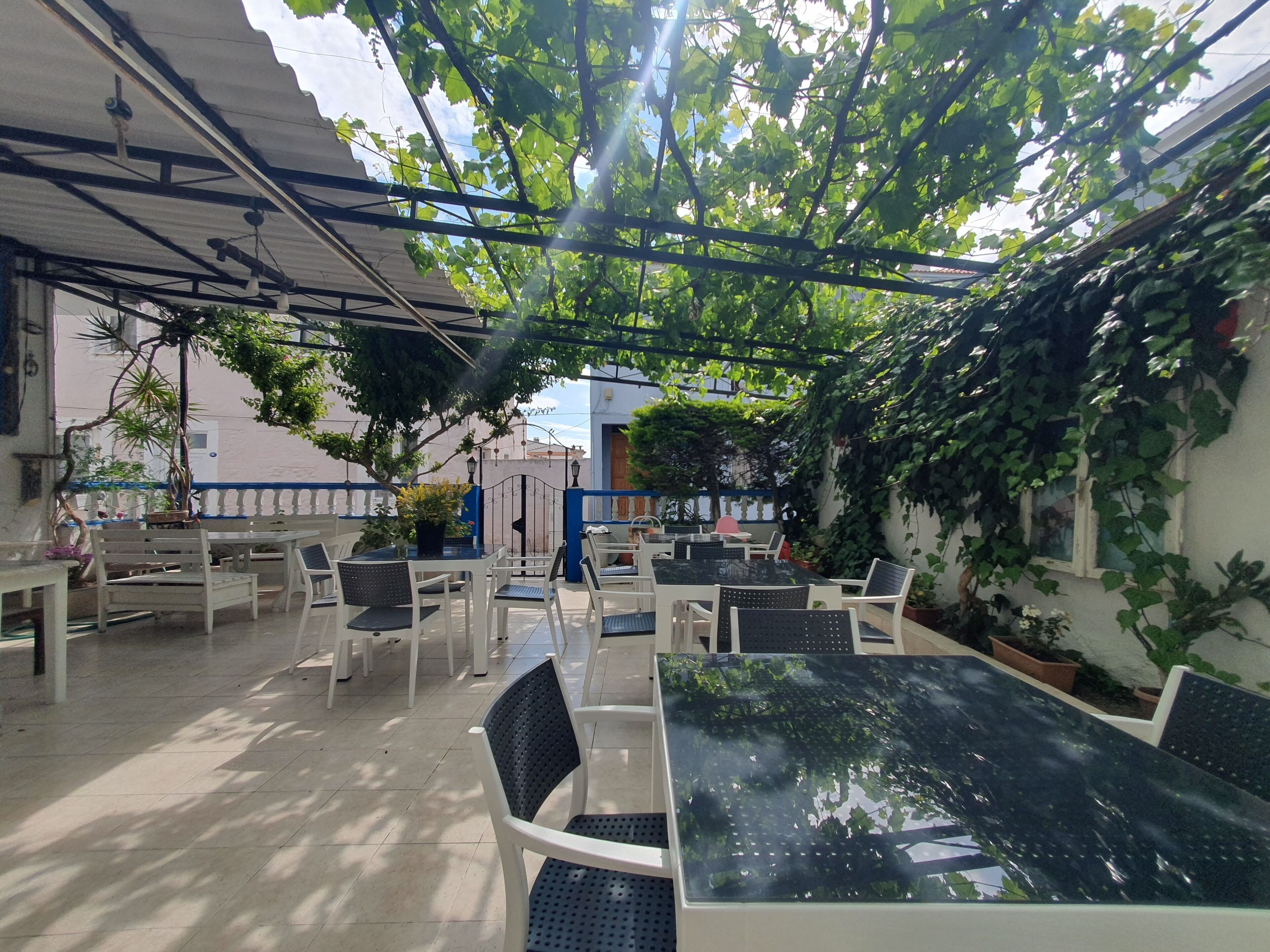 A covered outdoor seating area with tables and chairs, surrounded by greenery and a tiled floor.