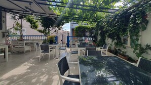 Een overdekt terras met tafels en stoelen, omgeven door groen en een betegelde vloer.