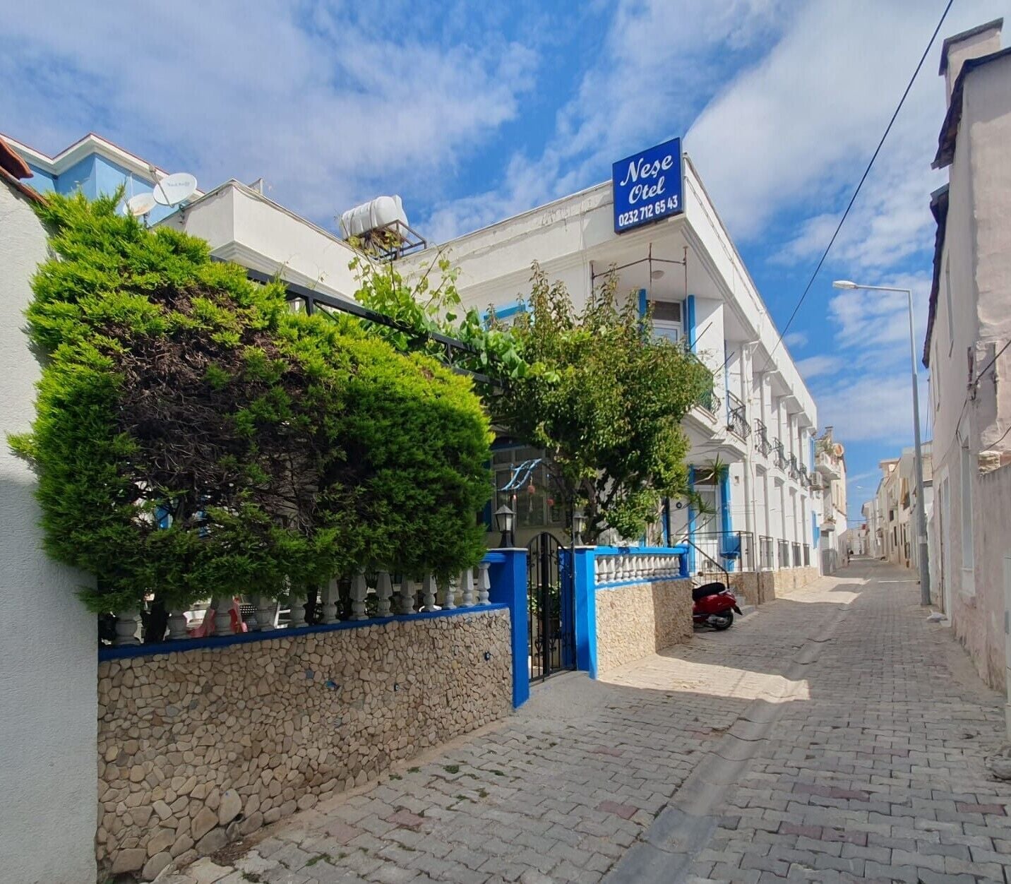 A narrow cobblestone street lined with white buildings, a blue sign for a hotel, and a red scooter parked on the side.