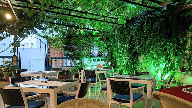 Een openluchtrestaurant met tafels en stoelen, omgeven door groen en een gebouw op de achtergrond.