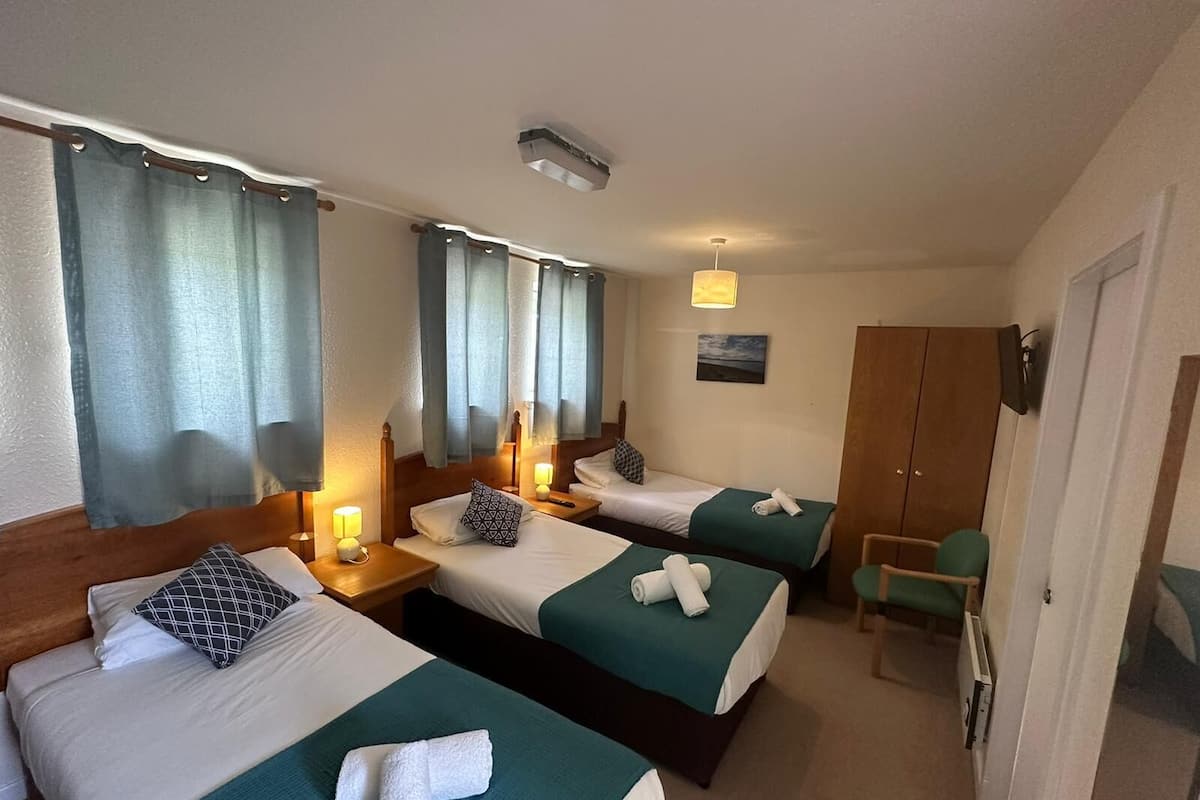 Une chambre d’hôtel avec trois lits simples, chacun recouvert d’une couverture verte et doté de draps blancs. On y trouve des lampes de chevet, un luminaire au plafond et une petite table avec une chaise. Une armoire en bois et une porte sont visibles à l’arrière-plan.