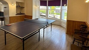 A room with a ping pong table, wooden chairs, a small kitchen area, and a window with a view of greenery. - Mackay's Spa Lodge Hotel (Strathpeffer)