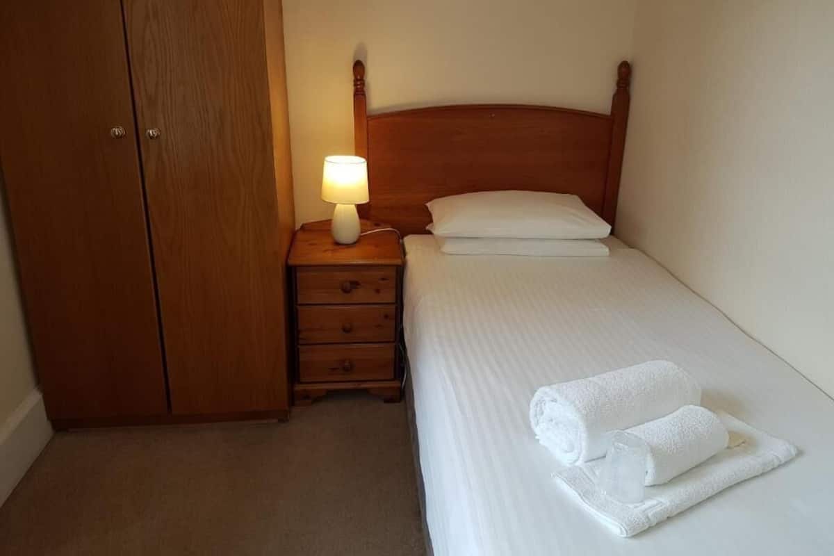 Une chambre avec une armoire en bois, une table de chevet avec une lampe, un lit bien fait avec du linge de lit et des serviettes blanches, et un mur sobre.