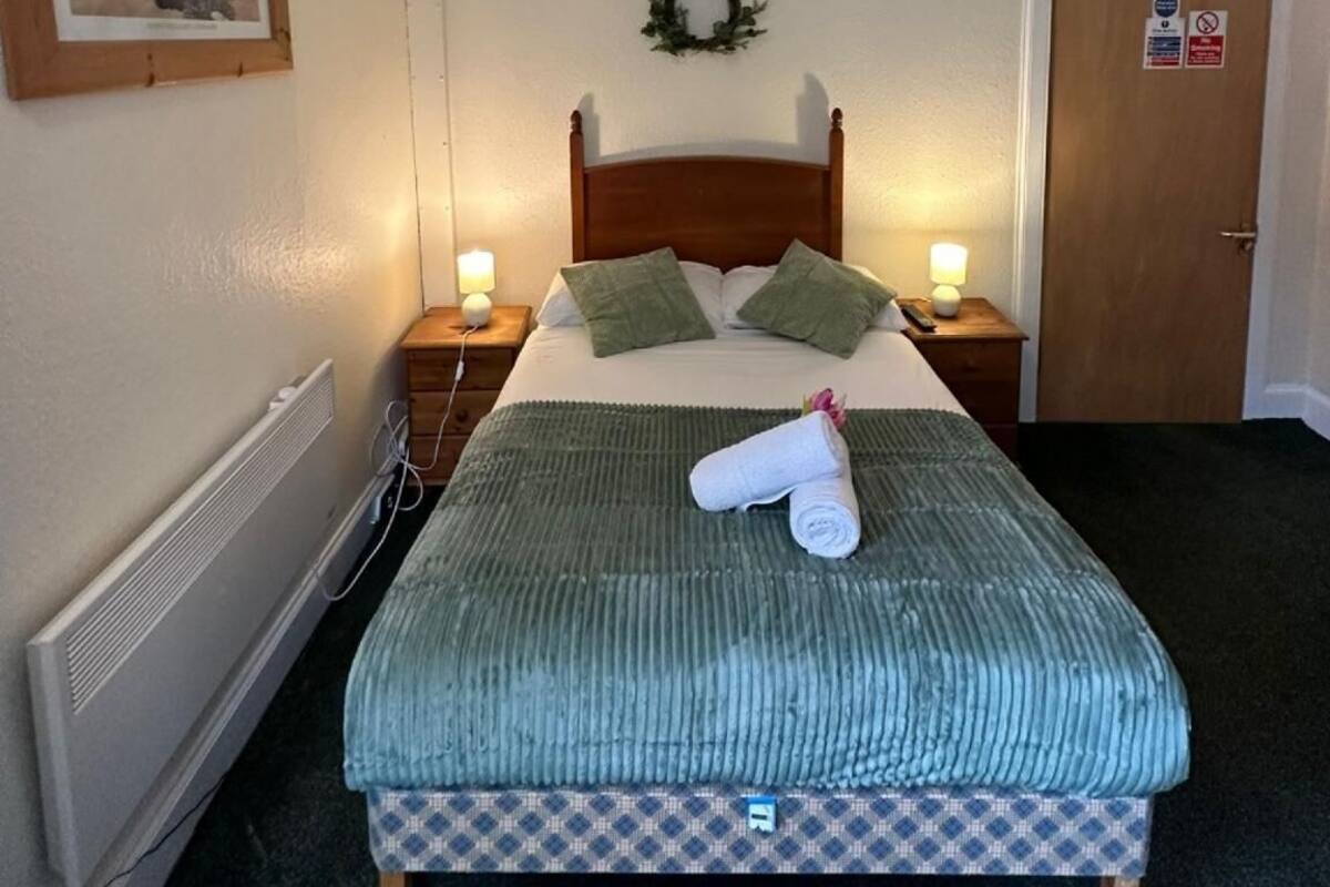 Un lit bien fait, recouvert d’une couverture verte, avec deux oreillers et une serviette enroulée posée sur le lit. Une petite table de chevet avec une lampe se trouve de chaque côté du lit. Un tableau encadré est accroché au mur, à gauche du lit.