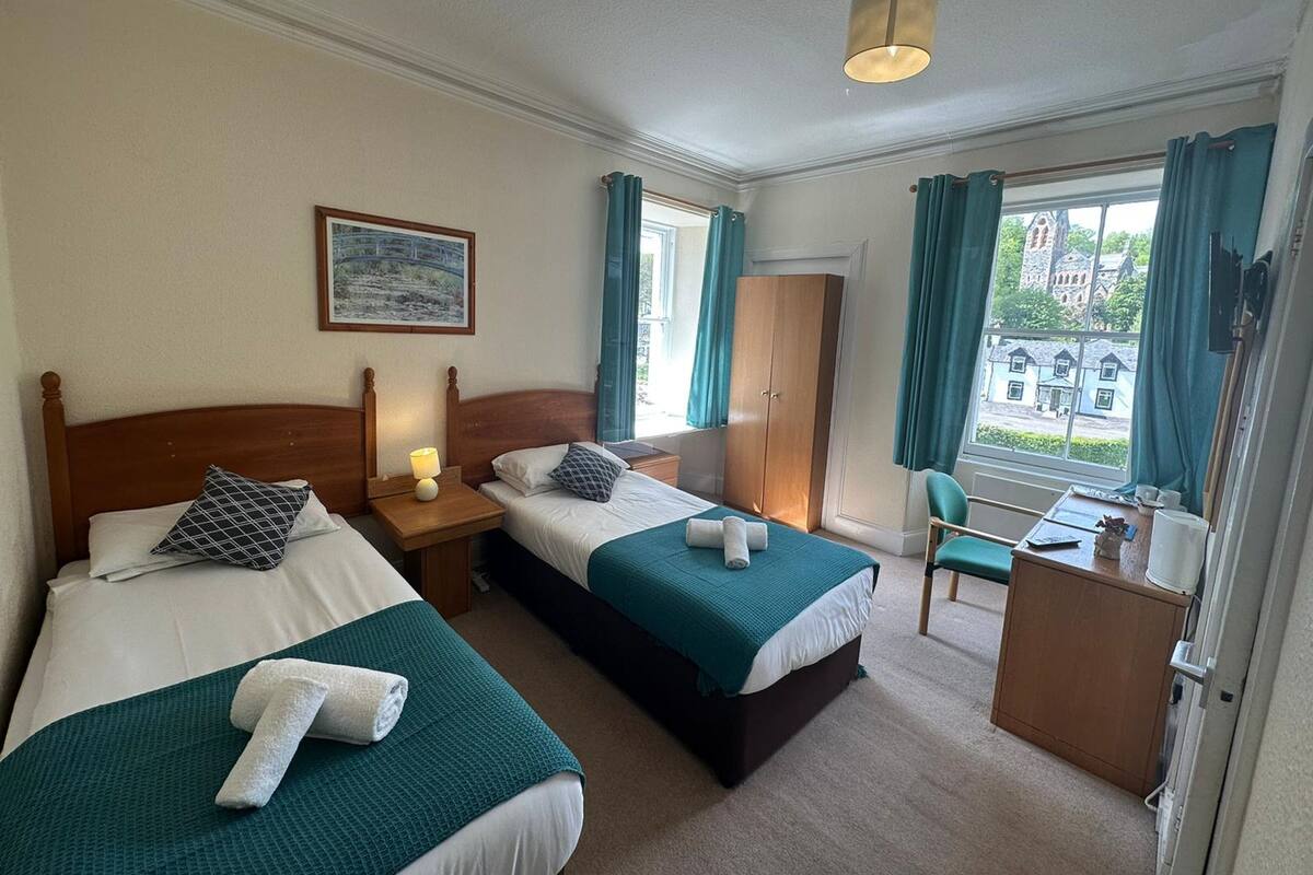 Une chambre d’hôtel avec deux lits simples, un bureau, une chaise, une armoire et une fenêtre avec des rideaux.