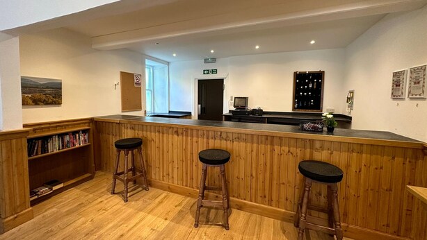 A reception area with a wooden counter, three bar stools, a bookshelf with books, a framed picture on the wall, and a small table with a camera and flowers. - Mackay's Spa Lodge Hotel (Strathpeffer)