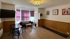A room with a wooden floor, a television mounted on the wall, a sofa, a table with chairs, and a chandelier. - Mackay's Spa Lodge Hotel (Strathpeffer)