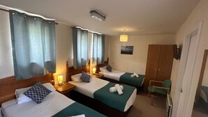A hotel room with three single beds, each with a pillow and a rolled towel. There is a wooden wardrobe, a desk with a chair, and a small table with a lamp. - Mackay's Spa Lodge Hotel (Strathpeffer)