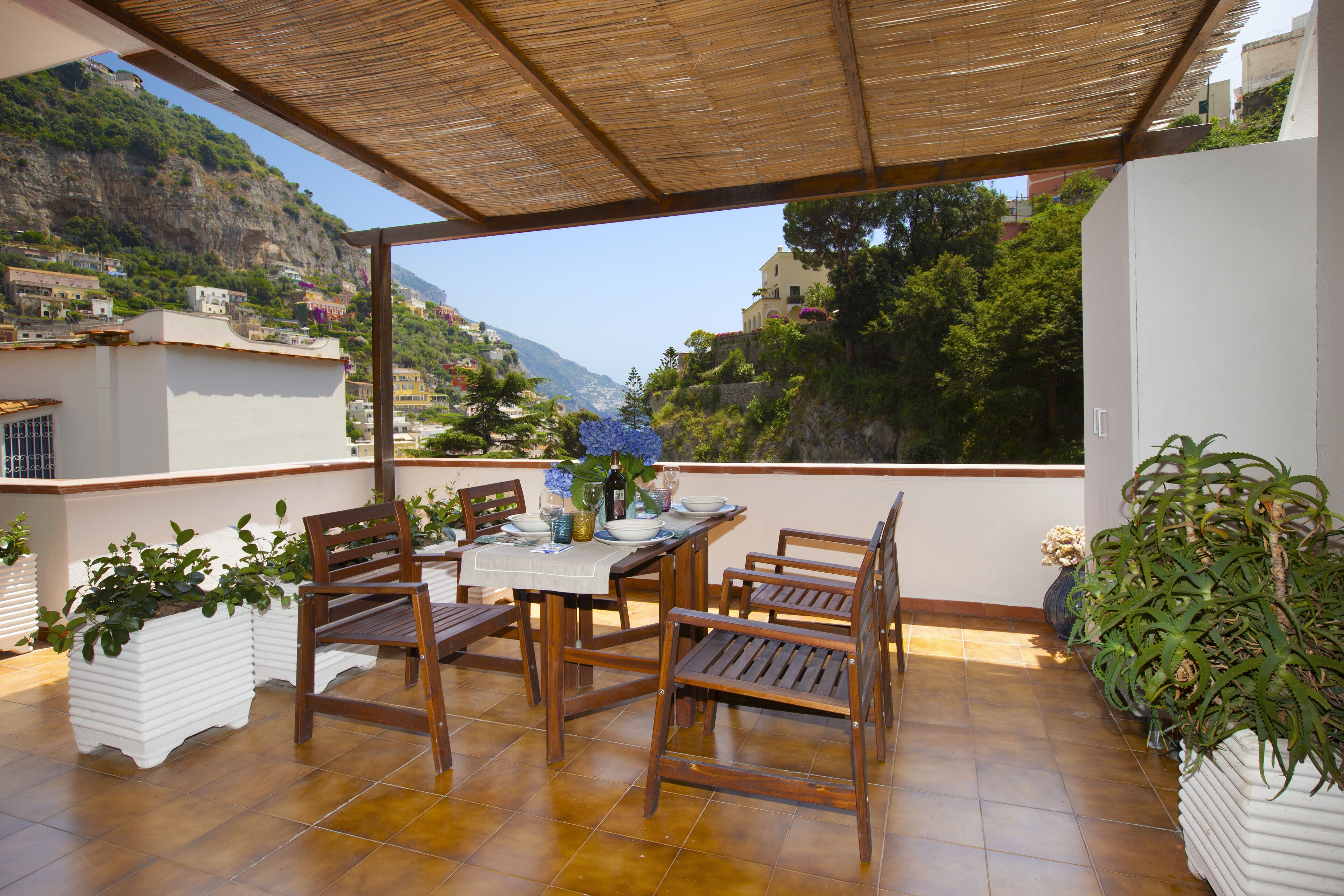 Un patio al aire libre cubierto con una mesa y sillas, rodeado de plantas en macetas y vistas a un paisaje montañoso.