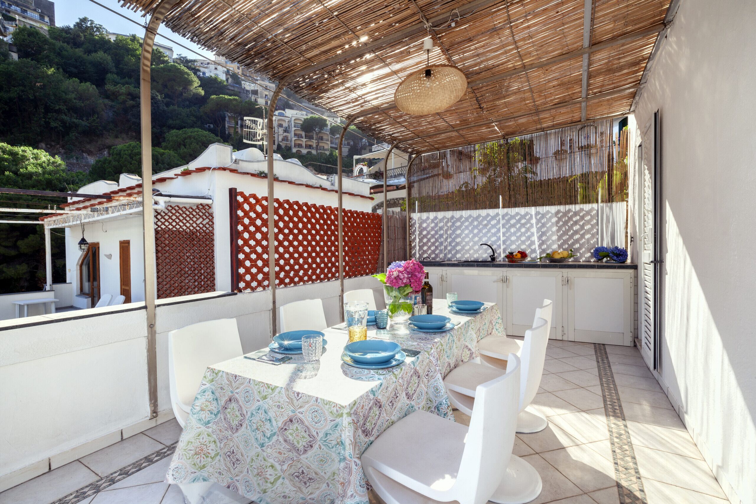A covered outdoor dining area with a table set for four, white chairs, and a kitchen area with a fruit bowl and utensils.