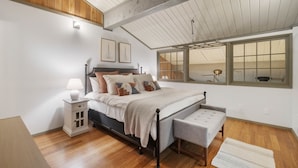 A bedroom with a sloped ceiling, a bed with a headboard, a bedside table with a lamp, a window with a view, and a bench.