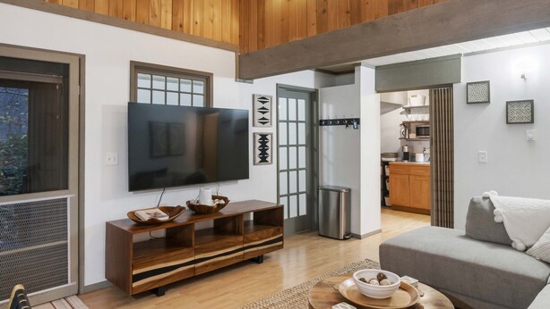 A spacious living area with a high ceiling, wooden walls, a large flat-screen TV, a grey sofa, and a round coffee table.