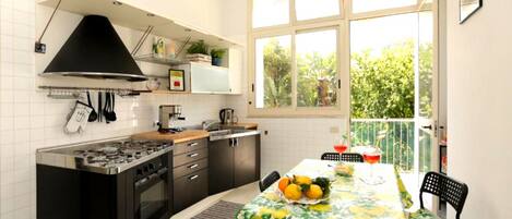 A compact kitchen with a gas stove, oven, and black range hood. A dining table with a floral tablecloth and chairs is set up near a large window with a view of greenery.