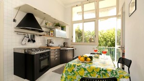 A kitchen with a stove, oven, and black cabinets. There is a dining table with a fruit bowl and a window overlooking greenery. - Estate4home - Casa Chiarina (Sorrento)