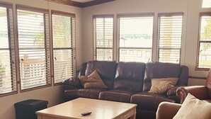 A living room with a brown leather sofa, a wooden coffee table, and large windows with blinds. - St. Johns River Retreat - CSC Unit 1 (Crescent City)