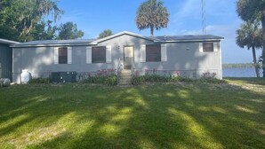 A single-story house with a front porch, a small garden with pink flowers, and a view of a lake. - St. Johns River Retreat - CSC Unit 1 (Crescent City)