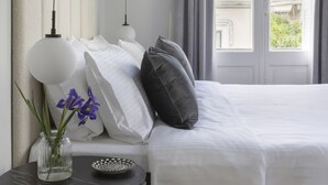 A neatly made bed with white linens and a gray pillow, a small round table with a vase of purple flowers, and a view of a building through a window with gray curtains. - Beautiful Acropolis Villa 3BR 9ppl 10 minutes Metro&Museum (Athina)