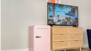 Un réfrigérateur rose, une commode en bois avec des tiroirs et un téléviseur diffusant une scène de plage.