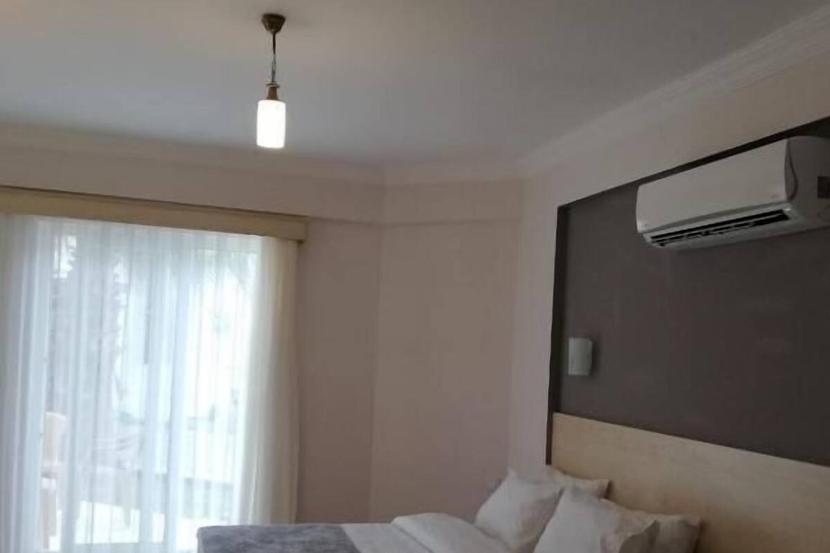 A hotel room with two beds, a large window with sheer curtains, an air conditioning unit, and a ceiling light.