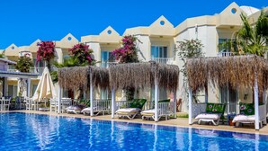 Uma área à beira da piscina com espreguiçadeiras e guarda-sóis, uma piscina azul cristalina e uma fileira de edifícios com estilo arquitetônico uniforme.