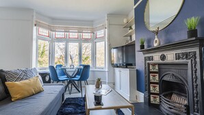 Ein Wohnzimmer mit Erkerfenster, Kamin, einer Couch, einem Esstisch und blauen Stühlen.