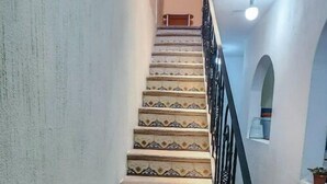 Un escalier aux marches richement décorées et à la rampe noire, menant à une pièce dont le sol est recouvert de carreaux et qui est équipée d’un ventilateur de plafond.