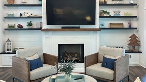 A modern living room with a large flat-screen TV, a fireplace, and two armchairs with cushions.