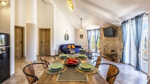 A dining area with a wooden table set for four, surrounded by chairs. A stone wall and a television are visible in the background. - Island Getaway Heritage House: Historical Villa with Heated Pool & Sea Views (Supetar)