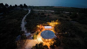 An aerial view of a circular stone building with a central courtyard, surrounded by a dirt path and sparse vegetation, overlooking a large body of water and distant city lights. - Island Getaway Heritage House: Historical Villa with Heated Pool & Sea Views (Supetar)