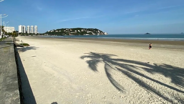 Ein weiter Sandstrand mit blauem Himmel, einer entfernten Hügelkette und einigen Gebäuden im Hintergrund.