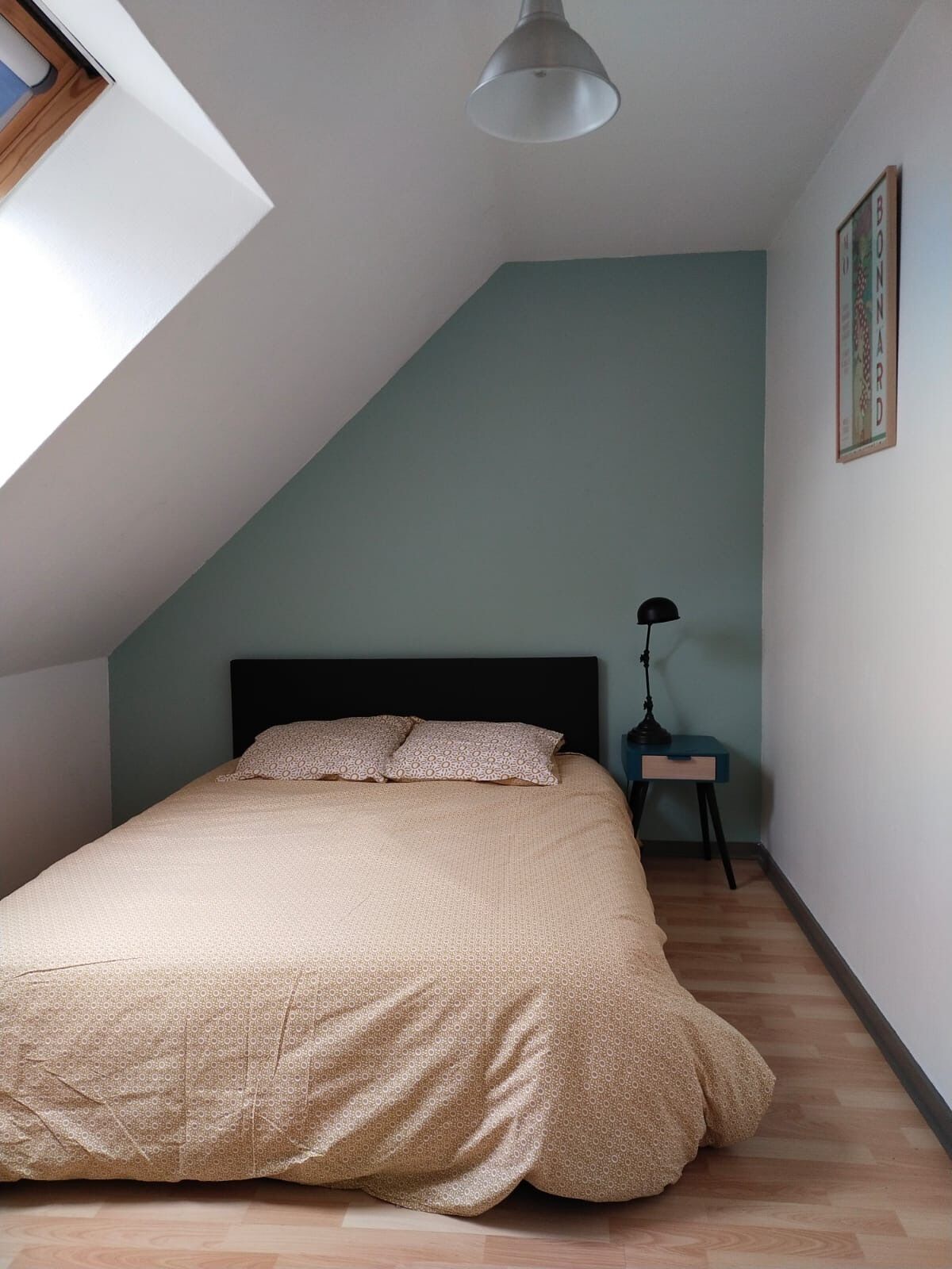 A bedroom with a sloped ceiling, a bed with patterned bedding, a nightstand with a lamp, and a framed picture on the wall.
