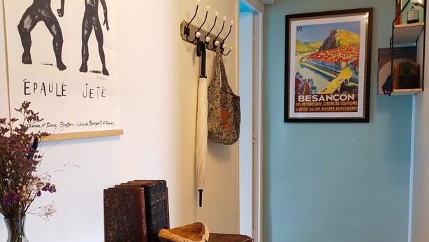 A hallway with a wooden dresser, a coat rack with a white umbrella, a framed poster, and a vase with flowers.