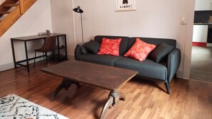 A living room with a dark sofa, a wooden coffee table, a small table with a chair, a lamp, and a poster on the wall.