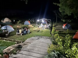 Un camping con varias tiendas de campaña, un espacio central para reunirse con mesas y sillas, y un sendero de piedra que lleva a las tiendas.