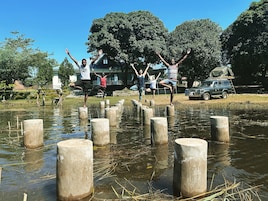 Un grupo de personas equilibrándose sobre postes de madera en una zona inundada, con árboles y un edificio al fondo.