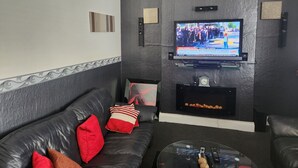 A modern living room with a leather sofa, a glass coffee table, a flat-screen TV mounted on a textured wall, and a fireplace below. - Inviting 3-bed House in Newcastle Upon Tyne (Newcastle-upon-Tyne)