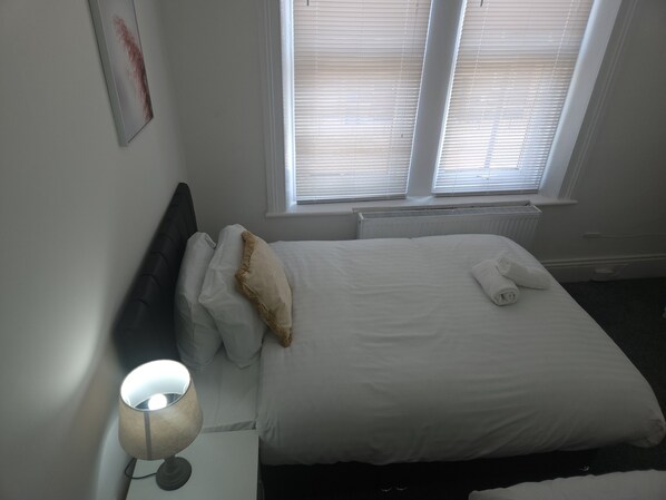 A neatly made bed with white linens and a beige throw pillow, a bedside lamp, and a radiator under a window with blinds.