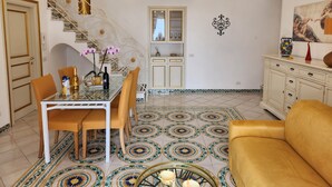 A dining area with a glass table, chairs, and a chandelier. There is a staircase with a decorative railing and a door with a phone.