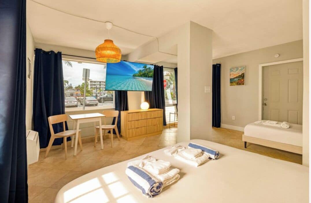 A hotel room with a bed, a desk, a chair, a TV, and a view of the beach.