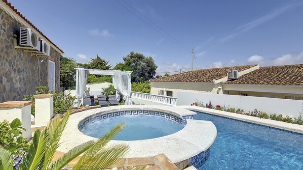 Ein Swimmingpool mit einem runden Whirlpool, umgeben von einer weißen Pergola, und ein Steinhaus mit einem Ziegeldach.
