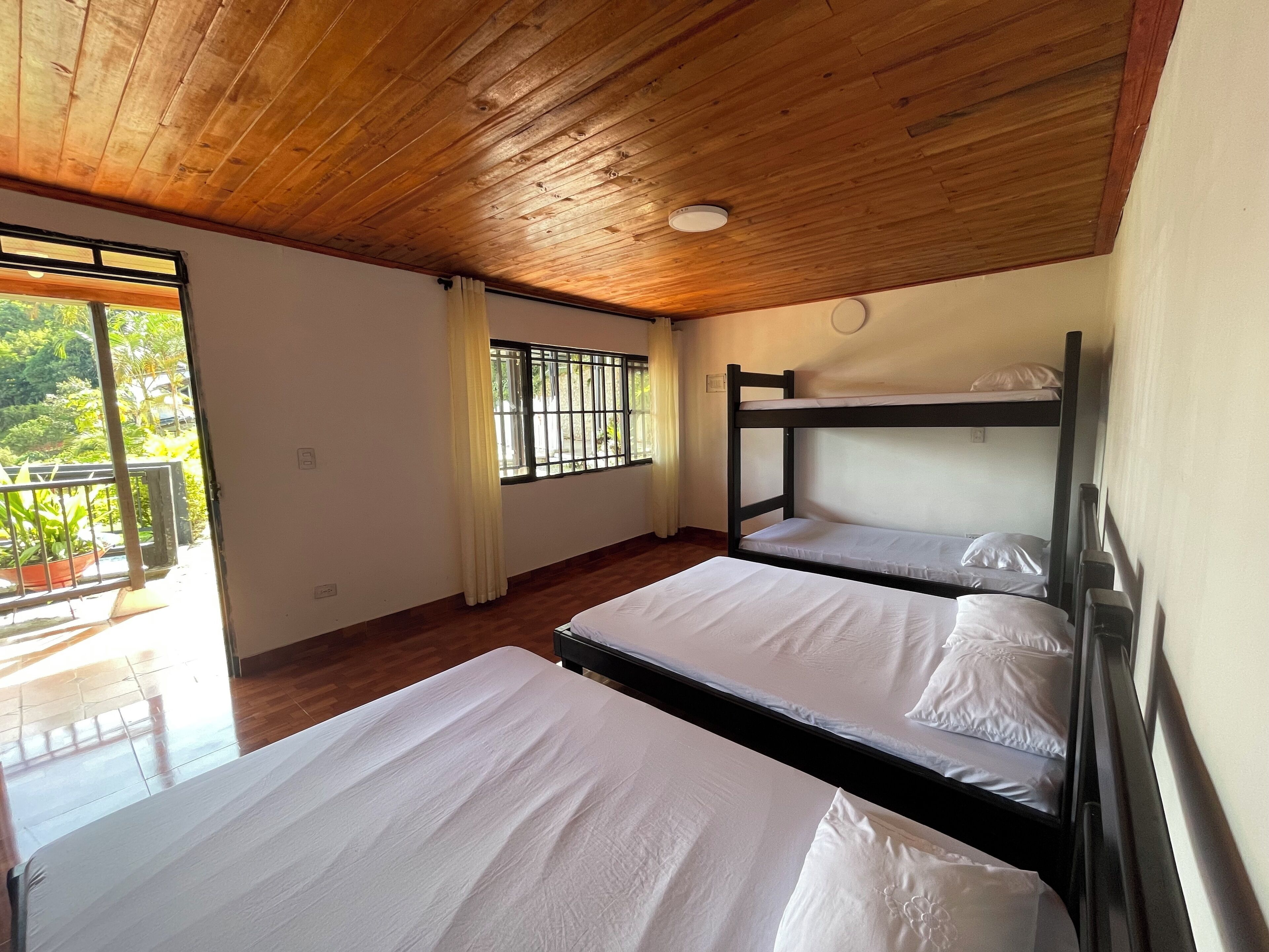 A room with wooden ceiling and bunk beds, a window with bars, and a view of greenery outside.