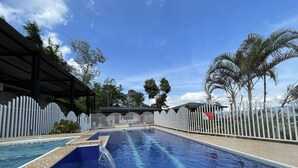 En swimmingpool med flisepanel, omgivet af et hvidt hegn, palmer og en klar blå himmel.