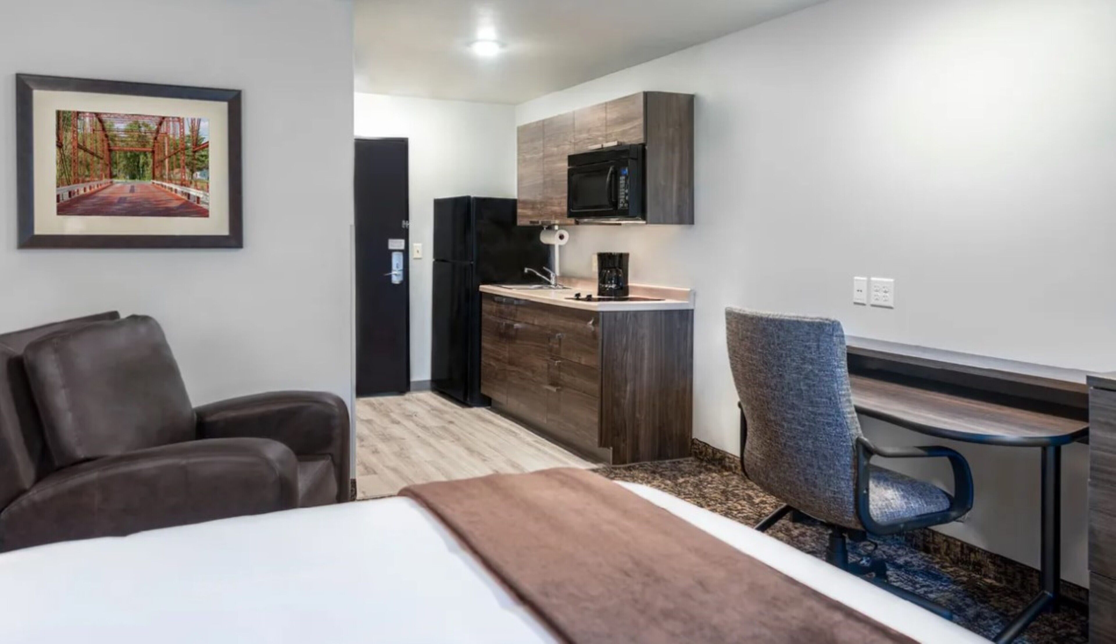 A compact hotel room with a bed, a desk, a chair, a kitchenette, and a framed picture on the wall.