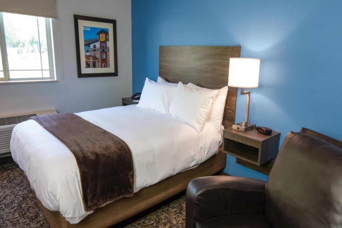 Habitación de hotel con cama, mesita de noche, silla, lámpara, cuadro en la pared y ventana con persianas.