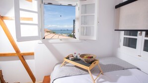 Quarto com uma janela grande, uma escada de madeira, uma cama e uma pequena mesa com uma cesta e garrafas.