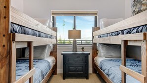 Quarto com duas camas beliche, uma secretária pequena com gavetas, uma luminária e uma janela com vista para árvores.